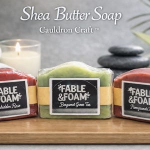 May include: Three shea butter soaps in red, green, and red hues, each wrapped in clear plastic and labeled with "Fable & Foam." The soaps are on a wooden surface, with a candle, towels, and stones in the background.