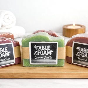 May include: Three rectangular bars of soap wrapped in clear plastic with brown paper bands. The labels read Fable & Foam with scents Forbidden Rose, Bergamot Green Tea, and Pomegranate Noir. The soaps are pink, green, and deep red in color.