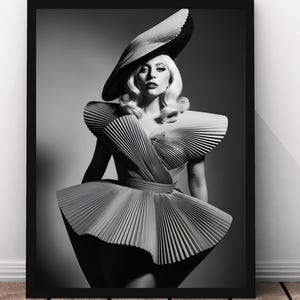 Może przedstawiać: Czarno-biała oprawiona grafika przedstawiająca portret osoby w dużym kapeluszu i plisowanej sukience. Zdjęcie studyjne ma neutralne tło. Osoba ma blond włosy i patrzy prosto w aparat.