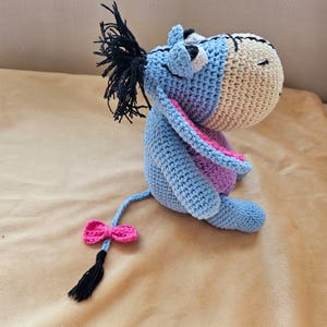 Può includere: Un peluche blu all'uncinetto che assomiglia a un asino con un fiocco rosa sulla coda. L'asino ha una criniera nera e un orecchio rosa e viola.