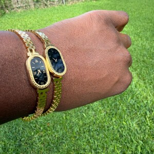 May include: Two gold-toned wristwatches with black faces and Roman numeral hour markers. The watches have an oval face and are on a gold chain-link bracelet. The watches are on a person's wrist, with a green grass background.