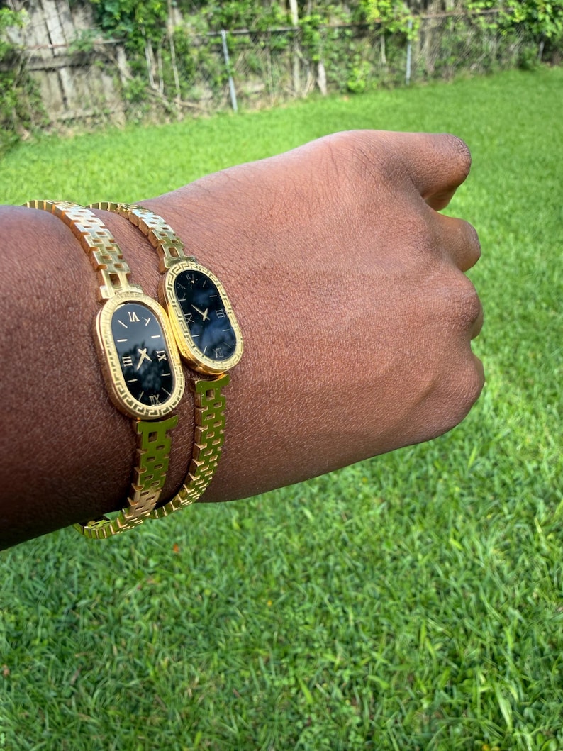 May include: Two gold-toned wristwatches with black faces and Roman numeral hour markers. The watches have an oval face and are on a gold chain-link bracelet. The watches are on a person's wrist, with a green grass background.