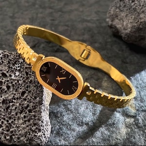 May include: Gold-toned wristwatch with a black oval face and Roman numeral hour markers. The watch features a gold bracelet-style band with a clasp closure. The watch is displayed on a textured gray surface.