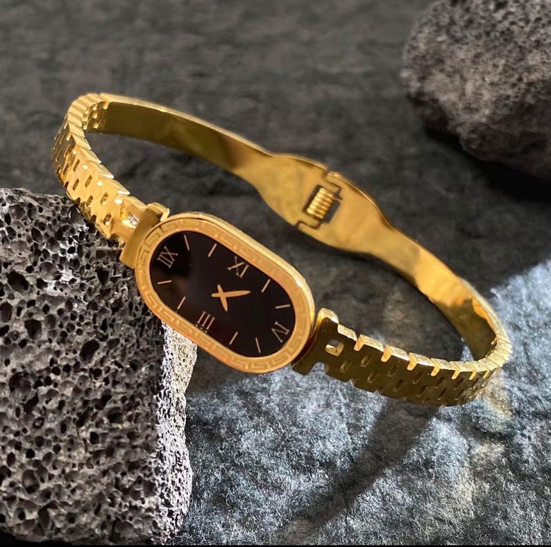 May include: Gold-toned wristwatch with a black oval face and Roman numeral hour markers. The watch features a gold bracelet-style band with a clasp closure. The watch is displayed on a textured gray surface.