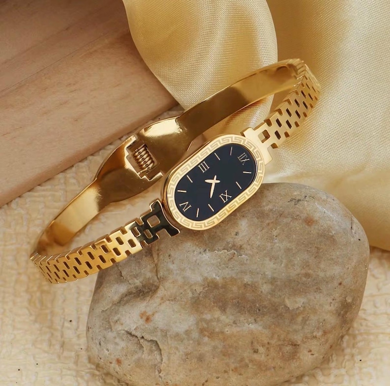 May include: Gold-toned bracelet watch with a black oval face and Roman numeral hour markers. The watch features a gold band and a clasp closure. The watch is displayed on a stone.