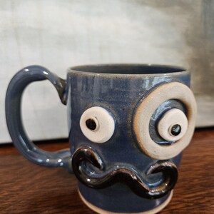 May include: A blue ceramic mug featuring a sculpted face with two mismatched white eyes, black pupils, and a prominent dark brown mustache. The mug has a curved handle on the left side and a smooth, glossy finish over the blue glazed surface.