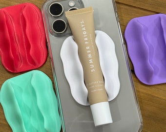 Puede incluir: Una funda de teléfono transparente con un agarre de teléfono blanco y un tubo de bálsamo labial Summer Fridays. El tubo está etiquetado como "Lip Butter Balm" y "Summer Fridays". La funda del teléfono está sobre una superficie de madera con tres agarres de teléfono de colores.