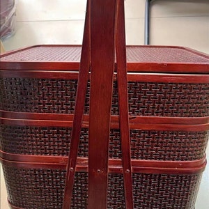 May include: A three-tiered, red wooden basket with a woven pattern and a long handle. The basket is made of bamboo and has a traditional Chinese design.