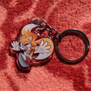 May include: A clear acrylic keychain featuring a cartoon character with a yellow and orange tail, white gloves, and a red shoe. The character is running and smiling. The keychain is attached to a silver metal ring.