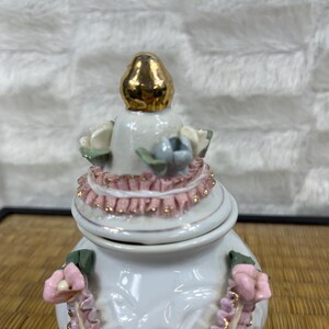 Puede incluir: Una caja de baratijas de cer&aacute;mica blanca con tapa dorada. La caja est&aacute; decorada con detalles florales rosas y blancos y un ribete rosa con volantes. La tapa est&aacute; rematada con un adorno dorado.