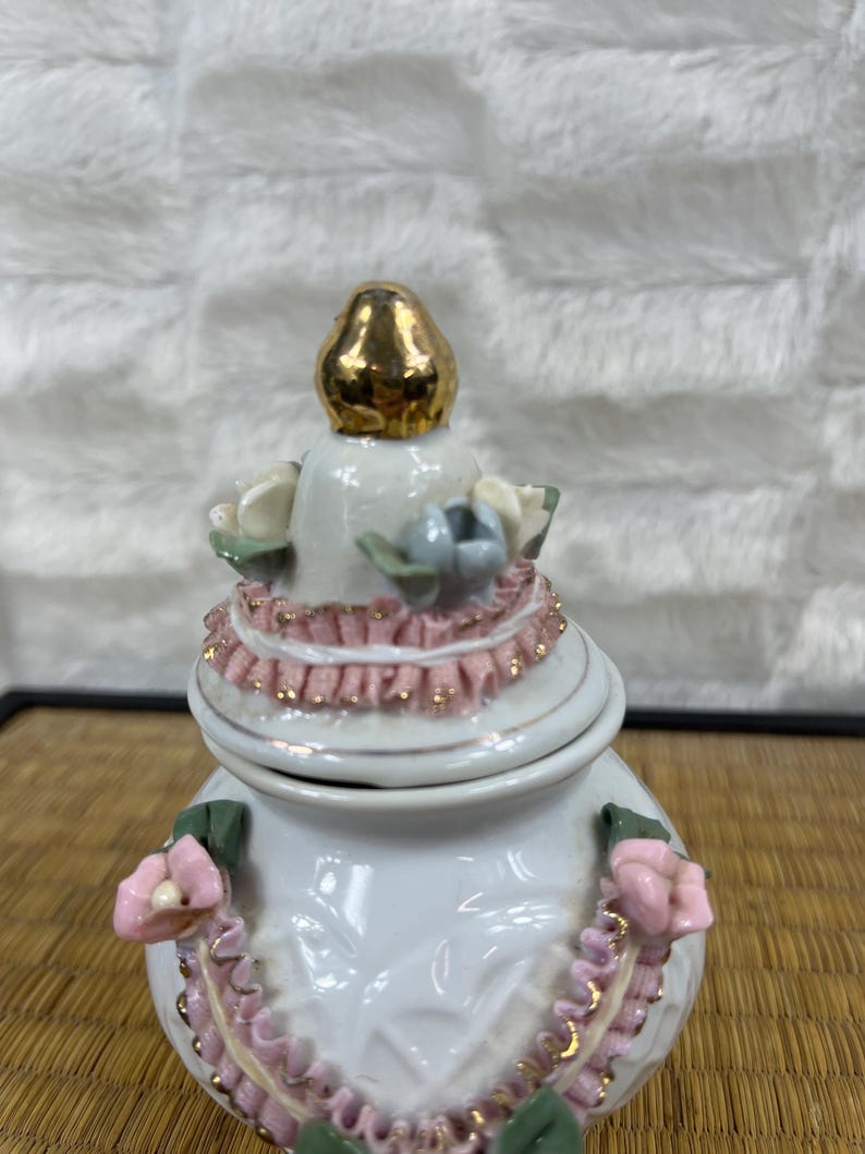 Puede incluir: Una caja de baratijas de cer&aacute;mica blanca con tapa dorada. La caja est&aacute; decorada con detalles florales rosas y blancos y un ribete rosa con volantes. La tapa est&aacute; rematada con un adorno dorado.