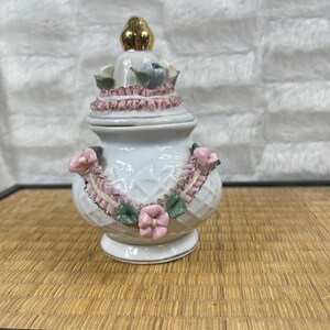 Puede incluir: Un joyero de cer&aacute;mica blanca con un remate dorado. El joyero est&aacute; decorado con detalles florales rosas y verdes y un ribete rosa en forma de cinta. El joyero tiene un patr&oacute;n de diamantes texturizado.