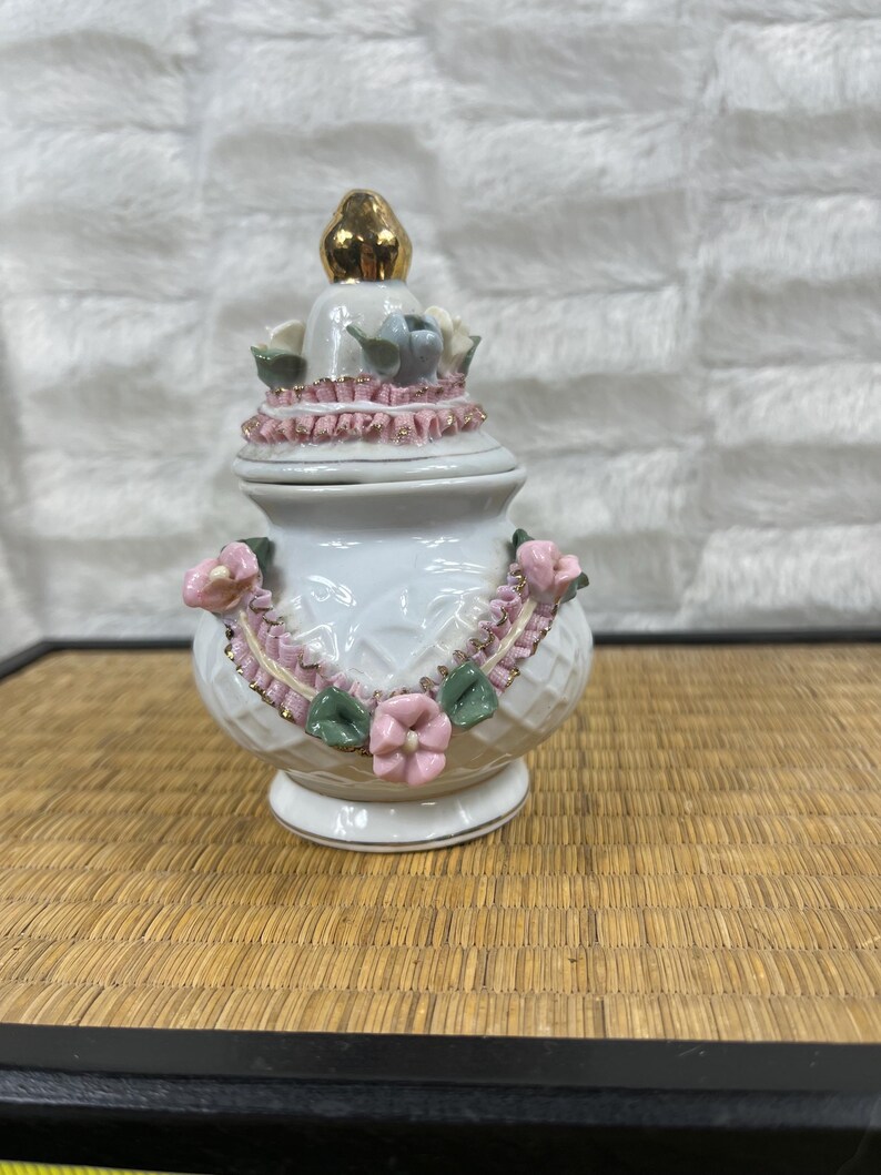 Puede incluir: Un joyero de cer&aacute;mica blanca con un remate dorado. El joyero est&aacute; decorado con detalles florales rosas y verdes y un ribete rosa en forma de cinta. El joyero tiene un patr&oacute;n de diamantes texturizado.