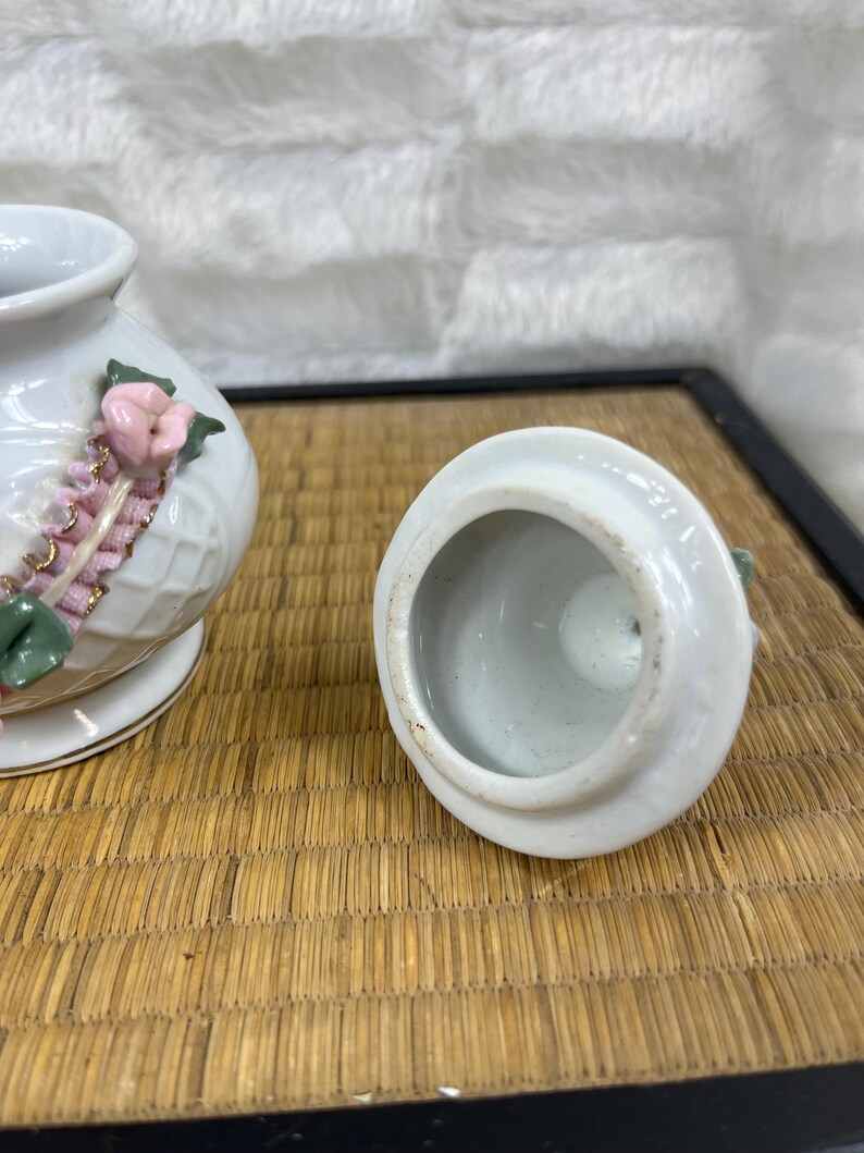 Puede incluir: Tarro de cer&aacute;mica blanca con tapa, decorado con detalles florales rosas y ribete dorado. El tarro tiene un patr&oacute;n texturizado, similar a un tejido de mimbre. La tapa est&aacute; quitada.