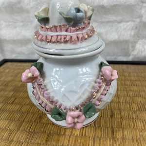 Puede incluir: Un tarro de cer&aacute;mica blanco con tapa, adornado con adornos rosas y dorados y detalles florales. El tarro tiene un remate dorado y est&aacute; decorado con flores rosas y hojas verdes. El tarro mide aproximadamente 13 cm de alto.