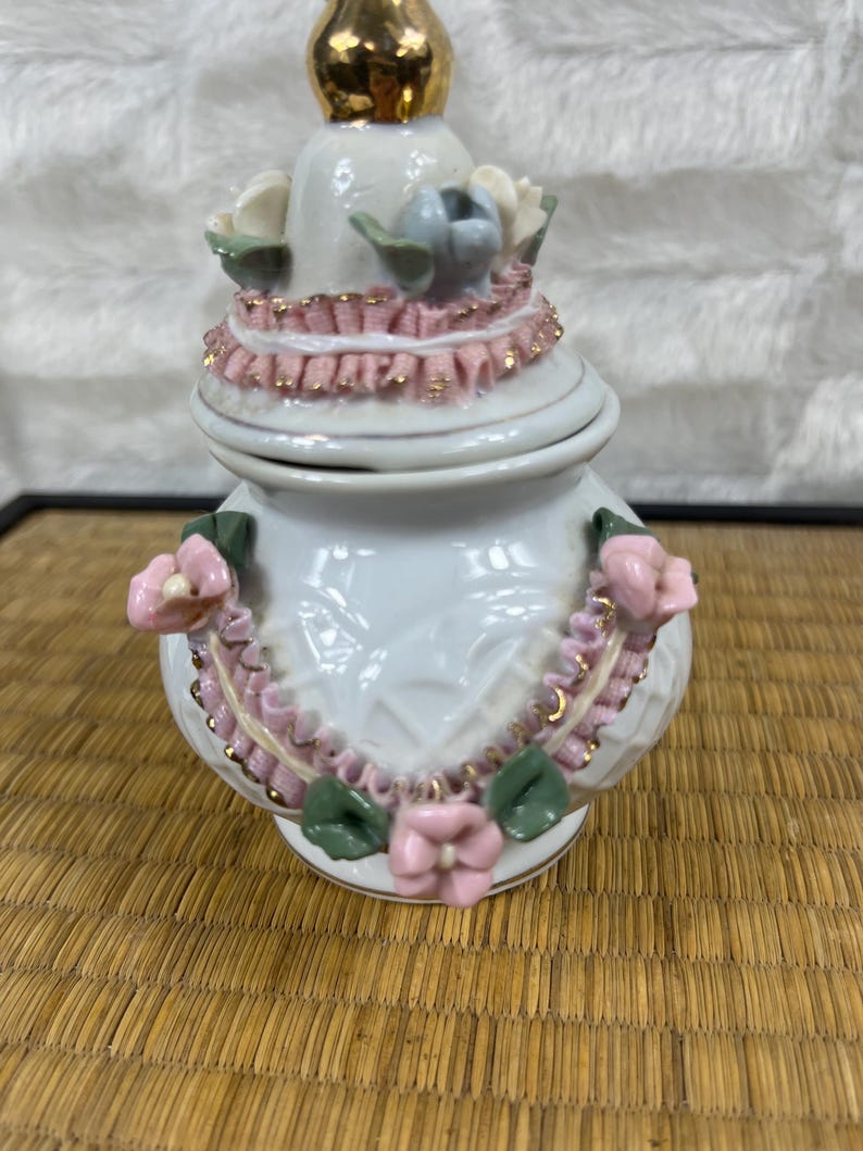Puede incluir: Un tarro de cer&aacute;mica blanco con tapa, adornado con adornos rosas y dorados y detalles florales. El tarro tiene un remate dorado y est&aacute; decorado con flores rosas y hojas verdes. El tarro mide aproximadamente 13 cm de alto.