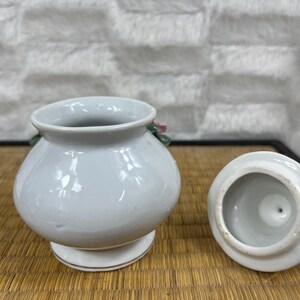 Puede incluir: Un peque&ntilde;o tarro de cer&aacute;mica blanca con tapa, colocado sobre una superficie tejida. El tarro tiene un cuerpo redondeado, una base de pedestal y un acento floral. La tapa est&aacute; a un lado, revelando el interior. El tarro probablemente se usa para almacenamiento o decoraci&oacute;n.