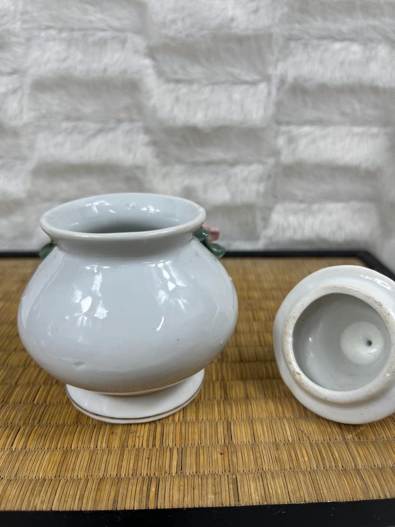 Puede incluir: Un peque&ntilde;o tarro de cer&aacute;mica blanca con tapa, colocado sobre una superficie tejida. El tarro tiene un cuerpo redondeado, una base de pedestal y un acento floral. La tapa est&aacute; a un lado, revelando el interior. El tarro probablemente se usa para almacenamiento o decoraci&oacute;n.