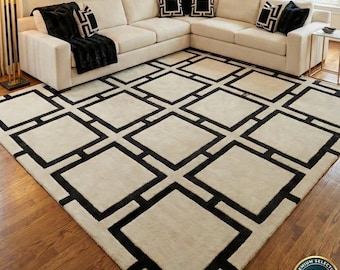 Può includere: Un grande tappeto con base color crema caratterizzato da un audace motivo geometrico a griglia nera composto da quadrati interconnessi. Il tappeto è posizionato su un pavimento in legno davanti a un grande divano angolare color crema con cuscini neri.