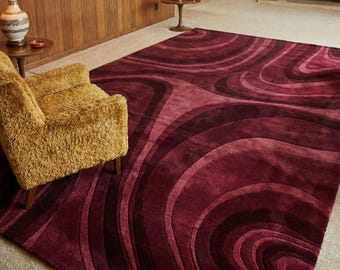 May include: A large area rug featuring a deep burgundy and maroon color palette with a repeating pattern of concentric, flowing, wave-like curves. The rug has a plush, textured pile that creates subtle shadows and highlights across the surface.