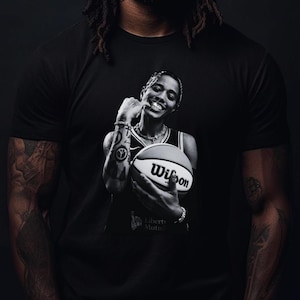 May include: A black short-sleeved t-shirt featuring a graphic print of a person holding a Wilson basketball. The graphic shows a person with tattoos on their arm, wearing a jersey and jewelry, smiling while holding the ball. The shirt is solid black.