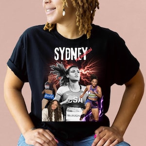 May include: Black t-shirt featuring a graphic print of Sydney McLaughlin, an American track and field athlete. The graphic includes the text "Sydney McLaughlin" and "USA".