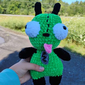 May include: A handmade green plush toy with large white eyes, black antennae, and black limbs. The toy has a pink tongue and a gray patch on its chest. The plushie is made of soft, textured yarn.