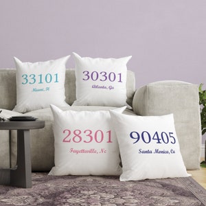 May include: Four white throw pillows with different city names and zip codes printed on them. The pillows are arranged on a light gray couch. The pillows are decorated with the following text: 33101 Miami, FL, 30301 Atlanta, GA, 28301 Fayetteville, NC, 90405 Santa Monica, CA.