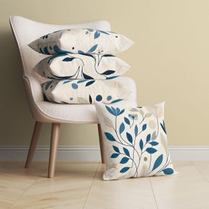 May include: Four decorative pillows with floral patterns in shades of blue, gray, and cream. Three pillows are stacked on a white chair, while the fourth rests on the floor. The pillows feature a botanical design.