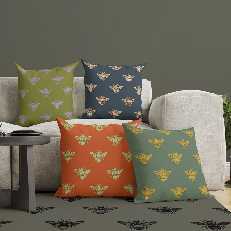 Puede incluir: Cuatro cojines cuadrados decorativos con un patr&oacute;n de abejas repetido. Los cojines son en tonos de verde, naranja y azul, con las abejas en colores contrastantes. Est&aacute;n colocados sobre un sof&aacute; de color claro, con una alfombra a juego.