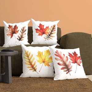 May include: Four square white throw pillows featuring watercolor illustrations of autumn leaves. Each pillow displays a unique arrangement of maple and oak leaves in shades of red, orange, yellow, and brown, set against a plain white background.