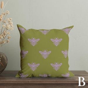 Puede incluir: Coj&iacute;n cuadrado verde oliva con un patr&oacute;n repetido de abejas color lavanda. El coj&iacute;n est&aacute; sobre una superficie de madera. La letra "B" est&aacute; en la esquina inferior derecha.