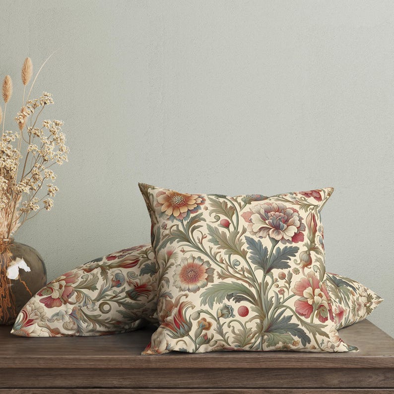 May include: A square throw pillow featuring a cream-colored background with a dense botanical pattern of large, multi-petaled flowers in shades of muted red, dusty pink, and soft orange, accented by intricate sage green and blue-grey foliage.
