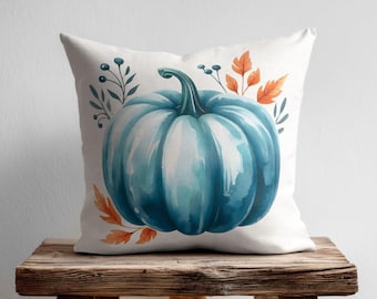 May include: A square throw pillow featuring a watercolor illustration of a large, vibrant teal pumpkin centered on a white background. The design is accented with delicate sprigs of dark blue berries and orange autumn leaves surrounding the pumpkin.