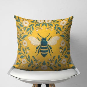 May include: Square decorative pillow with a yellow background featuring a large embroidered bee in teal and white. The bee is surrounded by a floral border with white flowers and teal leaves. The pillow is on a white chair.