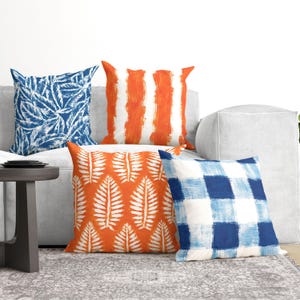 May include: Four square throw pillows featuring distinct patterns: one with blue and white leaves, one with orange and white vertical stripes, one with orange and white leaves, and one with a blue and white checkered pattern on a white sofa.
