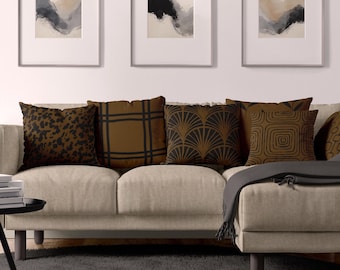 May include: A collection of square throw pillows arranged on a light beige sofa. The pillows feature various dark brown and black patterns, including animal print, grid lines, fan-shaped motifs, and concentric square designs in a cohesive color palette.