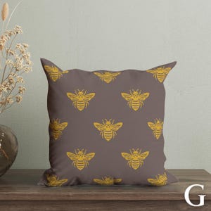 Puede incluir: Un coj&iacute;n decorativo cuadrado con un patr&oacute;n repetido de abejas amarillas sobre un fondo marr&oacute;n. El coj&iacute;n est&aacute; hecho de tela suave y est&aacute; dise&ntilde;ado para la decoraci&oacute;n del hogar.