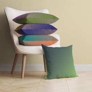 Peut inclure: Quatre coussins posés sur un fauteuil blanc. Les coussins sont de couleurs différentes, avec un effet dégradé. Le coussin du haut est vert et marron, le deuxième est violet et bleu, le troisième est turquoise et orange, et le dernier est orange uni. Un cinquième coussin est posé sur le sol, vert et bleu avec un effet dégradé.