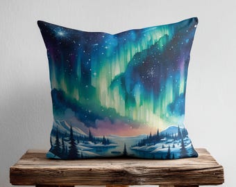 May include: A square throw cushion featuring a vibrant watercolour illustration of the aurora borealis in shades of teal, blue, and purple. The scene depicts a snowy landscape with dark silhouettes of pine trees and a starry night sky above.