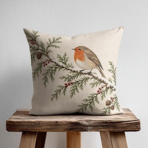 May include: A square throw pillow with a cream-colored fabric base featuring a detailed illustration of a robin perched on a green coniferous branch. The branch is adorned with small red berries and brown pinecones, creating a natural scene.