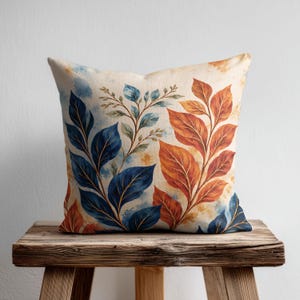 May include: A square scatter cushion featuring a watercolour-style print of botanical leaves. The design displays deep blue leaves on the left and warm orange-toned leaves on the right, set against a cream-coloured background with subtle paint splatters.