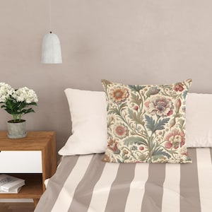 May include: A square throw pillow featuring a detailed botanical pattern with muted red, pink, and sage green flowers and swirling foliage on a cream background. The pillow is positioned on a bed with a light brown and white striped cover.