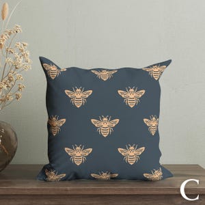 Puede incluir: Un coj&iacute;n decorativo cuadrado con un fondo azul oscuro y un patr&oacute;n repetido de abejas de color beige. El coj&iacute;n est&aacute; sobre una superficie de madera, con un jarr&oacute;n de flores secas en el fondo.