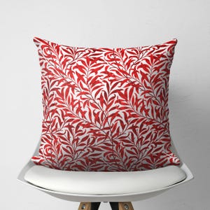 May include: Square throw pillow with a red and white botanical pattern. The pillow features a repeating design of red leaves and branches against a white background. The pillow is placed on a white chair.