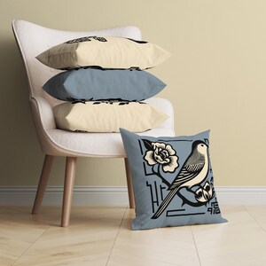 Puede incluir: Una almohada azul con una ilustración en blanco y negro de un pájaro posado en una rama con una flor. La almohada está sentada en una silla blanca con otras almohadas.