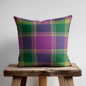 May include: A square throw pillow featuring a vibrant plaid pattern in shades of purple, green, and lime yellow. The design consists of intersecting horizontal and vertical lines forming a classic tartan grid across the entire surface of the pillow.