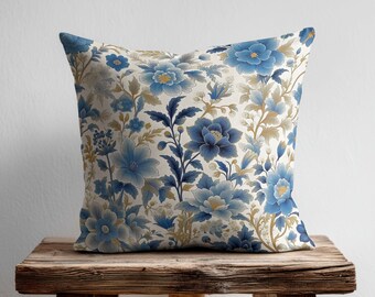 May include: A square throw pillow featuring a detailed botanical pattern with various shades of blue and muted gold flowers, leaves, and stems set against a light cream background. The design covers the entire front surface of the pillow.
