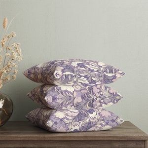 May include: Three decorative throw pillows with a floral pattern in shades of purple and cream. The pillows are stacked on top of each other on a wooden surface.