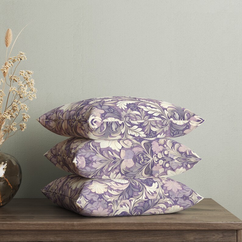 May include: Three decorative throw pillows with a floral pattern in shades of purple and cream. The pillows are stacked on top of each other on a wooden surface.